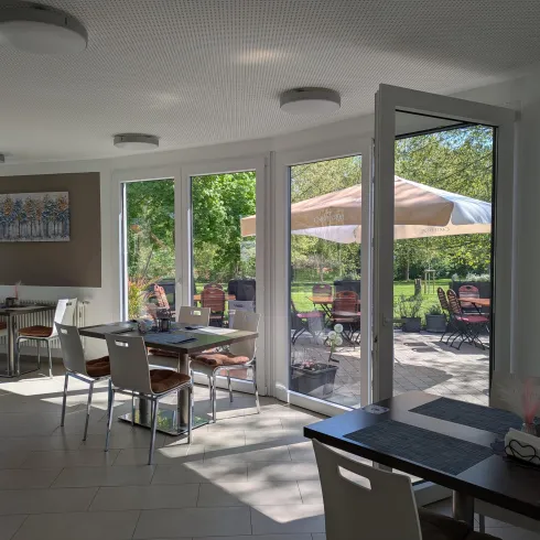 Ein helles und modernes Restaurant mit großen Fenstern und Blick auf den Außenbereich. Die Tische sind elegant gedeckt und umgeben von Pflanzen.