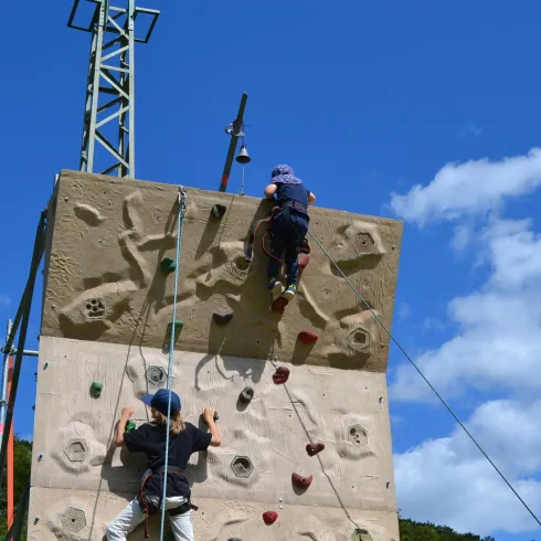 Zwei Kinder klettern an einer Kletterwand unter einem klaren, blauen Himmel. Die Wand ist mit bunten Griffen ausgestattet und bietet eine spannende Herausforderung.