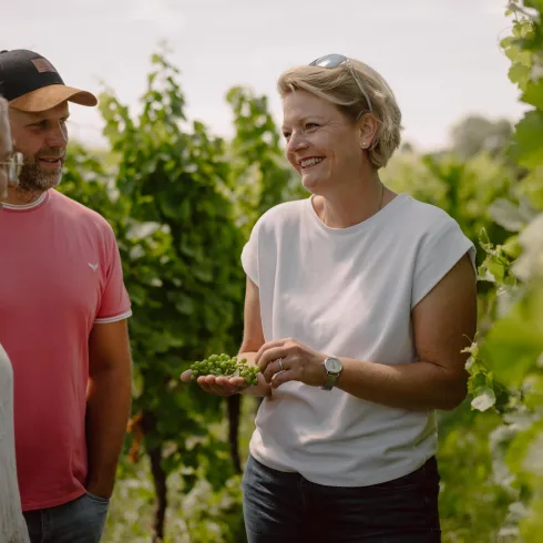 Eine Frau erklärt etwas in einem Weinberg, während sie mit zwei Personen spricht. Die Umgebung ist grün und üppig mit Weinreben.