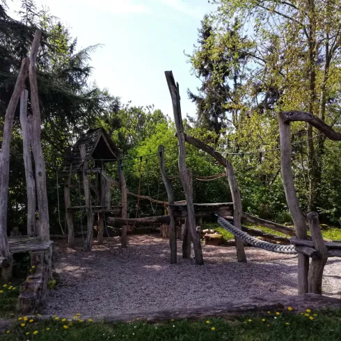 Ein Spielplatz aus Holz mit verschiedenen Kletterelementen. Umgeben von Bäumen und einer natürlichen Umgebung.
