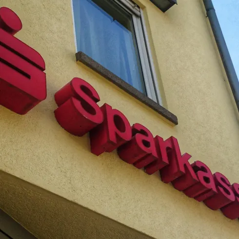 Ein rotes Schild mit dem Schriftzug "Sparkasse" an einer gelben Wand. Das Schild ist deutlich sichtbar und gut lesbar.