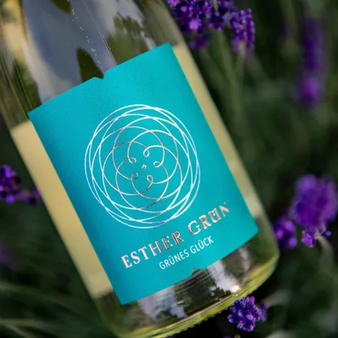 Eine Flasche Weißwein mit dem Etikett „Esther Grün – Grünes Glück“ steht vor lila Lavendelblüten. Der Hintergrund zeigt eine natürliche, blühende Umgebung.