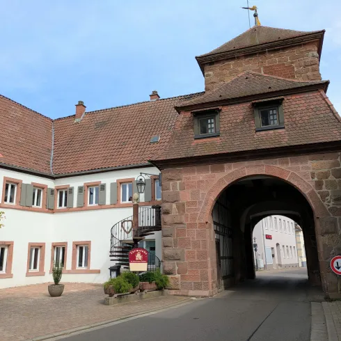 Ein historisches Tor mit einem roten Ziegeldach und angrenzenden Gebäuden. Die Straße führt durch das Tor in eine ruhige Umgebung.