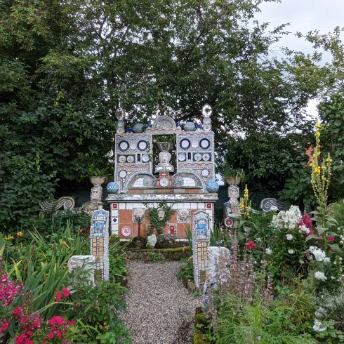 Ein bunter Garten mit ungewöhnlichen, kunstvoll gestalteten Skulpturen und vielen Blumen. Bäume und Sträucher umrahmen den Bereich und schaffen eine einladende Atmosphäre.
