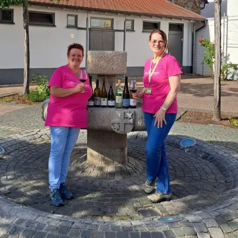 Zwei Frauen in pinken T-Shirts stehen an einem Brunnen und halten Weingläser. Hintergrund zeigt eine ruhige, gepflasterte Umgebung.