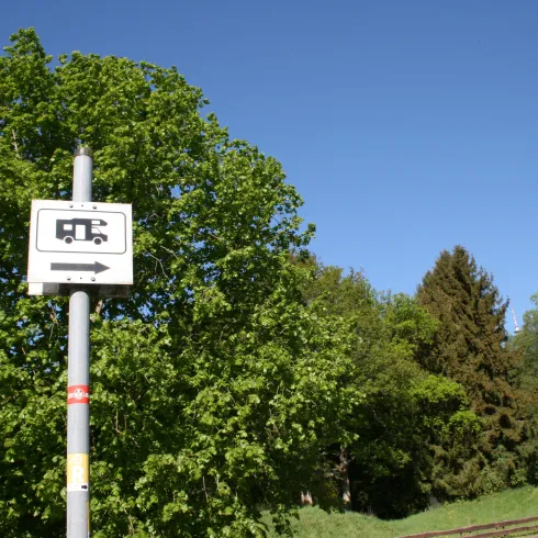 Ein Schild zeigt die Richtung zu einem Wohnmobilstellplatz. Im Hintergrund sind grüne Bäume und ein klarer blauer Himmel sichtbar.
