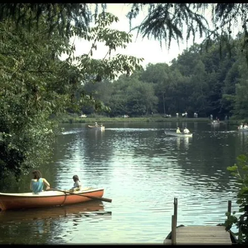 Ein ruhiger See umgeben von Bäumen, auf dem Boote fahren. Zwei Personen sitzen in einem orangefarbenen Boot und genießen die Natur.