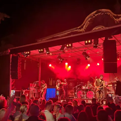 Ein Konzert unter den Sternen mit einer Band auf der Bühne. Die Menge genießt die Musik bei heller Beleuchtung.