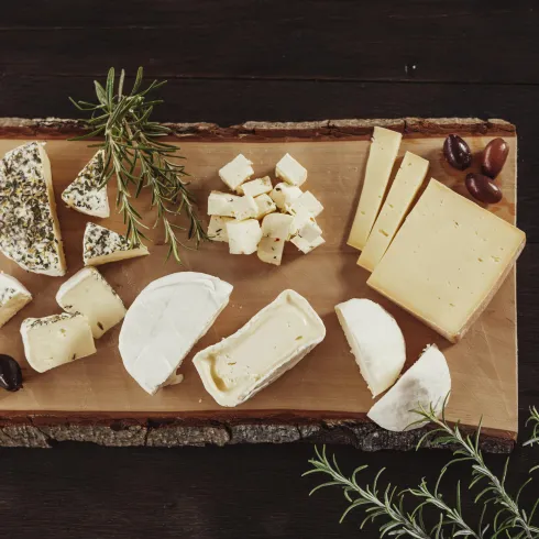 Eine Auswahl an verschiedenen Käsesorten auf einem Holzbrett. Neben dem Käse befinden sich einige Oliven und frische Kräuter.