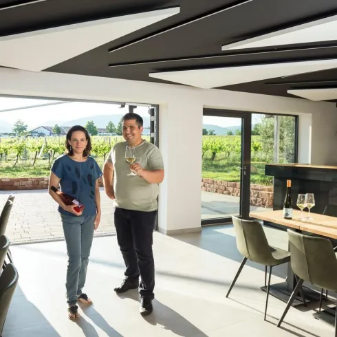 Ein Weinliebhaber zeigt stolz seine Weinsammlung in einem modernen Weingut. Im Hintergrund sind Weinberge und eine helle, einladende Atmosphäre zu sehen.