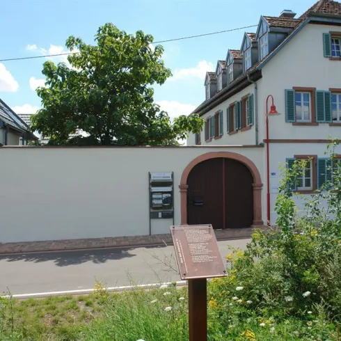 Ein historisches Gebäude mit einer rustikalen Fassade und Holzfenstern. Im Vordergrund gibt es eine informativen Tafel und etwas grünes Gras.