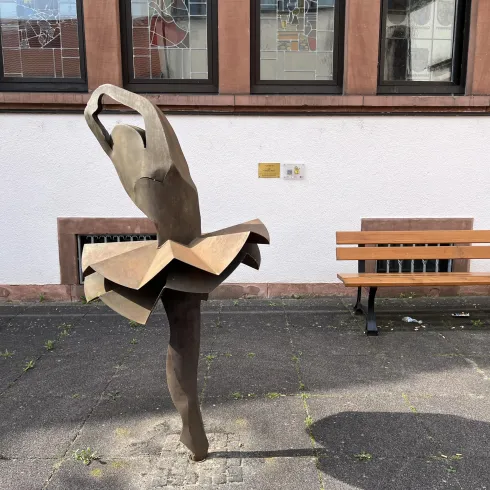 Eine bronze Farbe Skulptur einer Ballerina steht auf einem Pflaster. Im Hintergrund ist eine Bank und ein Fenster zu sehen.