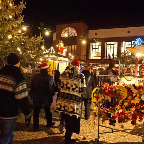 Ein festlicher Weihnachtsmarkt mit Lichtern und Ständen. Menschen in winterlicher Kleidung genießen die Atmosphäre und stöbern nach Geschenken.