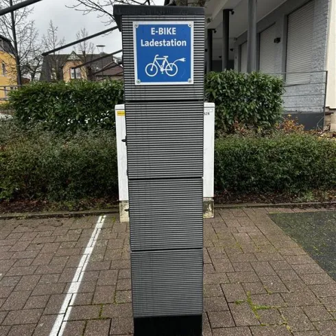 Eine E-Bike Ladestation steht in einem Straßeneck. Es ist bewölkt und die Umgebung besteht aus Bäumen und Gebäuden.