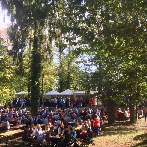 Eine belebte Outdoor-Veranstaltung mit vielen Menschen, die an Tischen sitzen. Im Hintergrund sind Zelte und Bäume zu sehen.