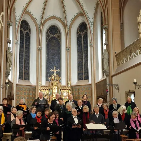Ein Chor steht in einer Kirche und singt. Im Hintergrund sind bunte Fenster und religiöse Statuen zu sehen.