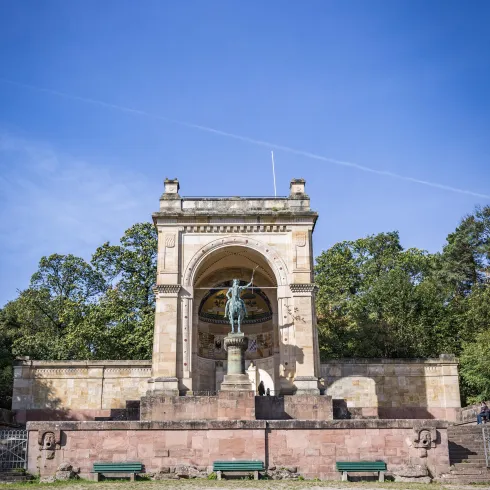 Ein historisches Denkmal mit einer Statue in einer monumentalen Nische. Umgeben von Bäumen und einem klaren, blauen Himmel.