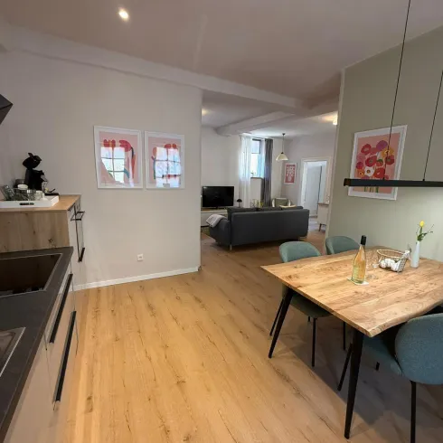 Eine moderne Küche mit Holzfußboden und einem Essbereich. Im Hintergrund ist ein gemütliches Wohnzimmer mit einem Sofa und Fernseher zu sehen.