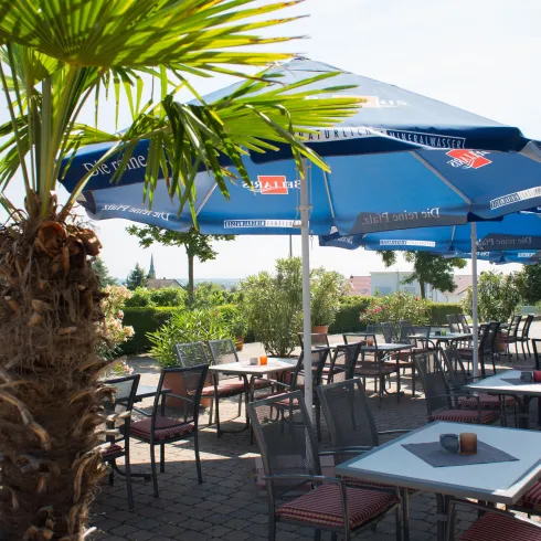 Ein gemütlicher Außenbereich eines Restaurants mit Sonnenschirmen und Tischen. Im Hintergrund sind Pflanzen und eine sonnige Atmosphäre sichtbar.