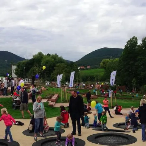Ein lebhafter Spielplatz mit vielen Kindern und Erwachsenen. Im Hintergrund sind grüne Hügel und Bäume zu sehen.
