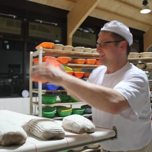 Ein Bäcker arbeitet in einer modernen Bäckerei und formt Teiglaibe. Im Hintergrund sind zahlreiche bunte Backformen zu sehen.