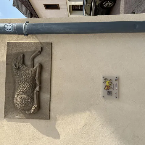 Ein Steinrelief eines Hundes befindet sich an einer Wand. Daneben ist ein Schild mit einem QR-Code angebracht.
