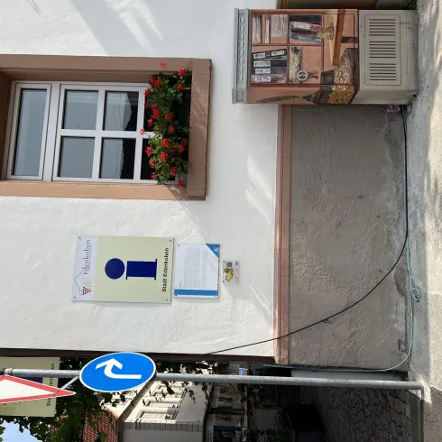 Ein Informationsschild an einer Wand mit einem Fenster und blühenden Pflanzen. In der Nähe steht ein Verkehrsschild für eine Einbahnstraße.