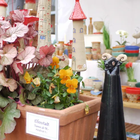 Ein bunter Blumentopf mit verschiedenen Pflanzen und einer kleinen roten Türmchen-Deko. Daneben stehen zwei schwarze Figuren, die Katzen darstellen.