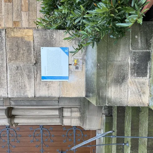 Ein historisches Gebäude mit einer Steinfassade und einer Treppe. An der Wand hängt ein Informationsschild, umgeben von Grünpflanzen.