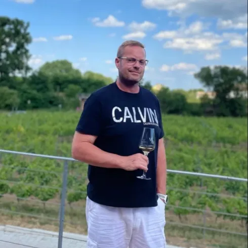 Ein Mann steht auf einem Balkon mit einem Glas Wein in der Hand. Im Hintergrund sind Weinberge und ein blauer Himmel mit einigen Wolken zu sehen.