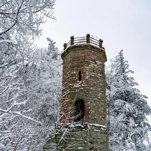 Ein alter Steinturm steht in einer schneebedeckten Landschaft. Um ihn herum sind winterliche Bäume mit Frost bedeckt.