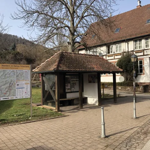 Eine Bushaltestelle mit einem Informationsschild und einem überdachten Wartebereich. Im Hintergrund ist ein traditionelles Gebäude und einige Bäume zu sehen.