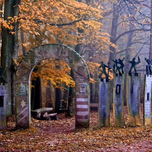 Ein mystischer Eingang zu einem Wald mit herbstlichen Bäumen. Das Tor ist kunstvoll gestaltet und von Nebel umhüllt.