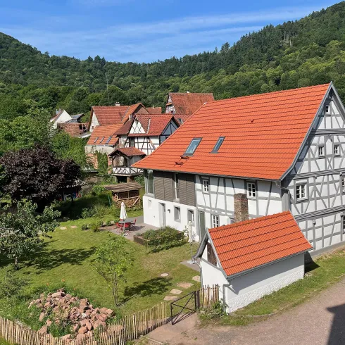 Eine malerische Landschaft mit typisch deutschen Fachwerkhäusern und einem großen Garten. Im Hintergrund sind bewaldete Hügel zu sehen und der Himmel ist klar und blau.