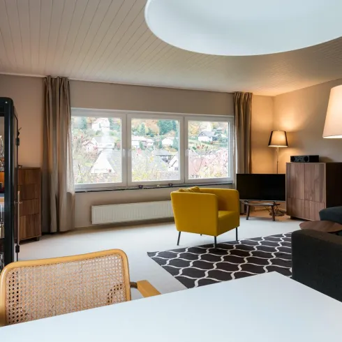 Ein modernes Wohnzimmer mit großen Fenstern und gemütlicher Einrichtung. Eine gelbe Couch und ein eleganter Essbereich ergänzen den Raum.