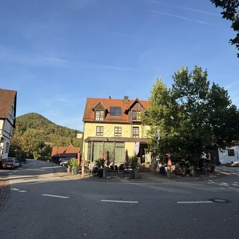 Ein charmantes Gebäude an einer Straßenecke mit roten Dächern und viel grünem Laub. Im Hintergrund sind sanfte Hügel und ein blauer Himmel sichtbar.