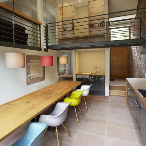 Eine moderne Küche mit einem langen Holztisch und bunten Stühlen. Die Räume sind hell und offen gestaltet, mit einem Balkon im oberen Bereich.