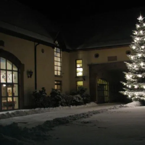 Ein festlich geschmückter Weihnachtsbaum steht vor einem Gebäude bei Nacht. Der Boden ist mit weißem Schnee bedeckt.