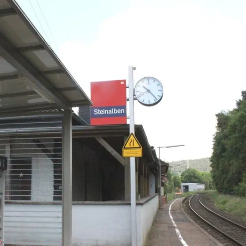 Der Bahnhof Steinraben mit einer Uhr und einem klaren Himmel. Die Gleise führen in die Ferne, umgeben von Bäumen.