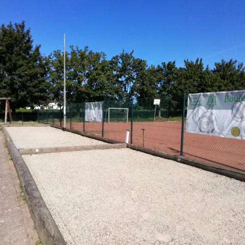 Ein Tennisplatz mit roten Belag und klar blauem Himmel. Im Hintergrund befinden sich Bäume und eine Sporteinrichtung.