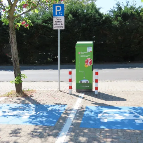 Die E-Tankstelle ist mit zwei blau makierten Symbolen versehen. Die E-Tanksäule ist von den Pfalzwerken. Man sieht im HIntergrund das die E-Tankstelle an der Straße liegt.