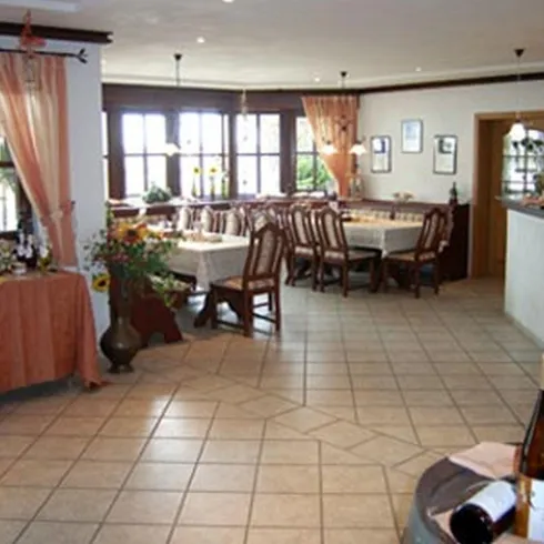 Ein Gastraum mit langen, bedeckten Tischen und einer Theke an der Wand. Die vielen Fenster sorgen für das Licht im Raum. Orange Details und Vorhänge sorgen für eine schöne Atmosphäre. 