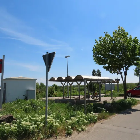 Man sieht ein gepflasterten Parkplatz der neben dem Bahnhof von Herxheim am Berg liegt. Der Parkplatz ist verschönert durch Pflanzenbeete und Bäume.