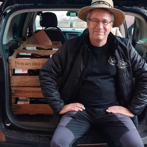 Ein Mann sitzt am Heck seines Autos, das mit Holzkisten gefüllt ist. Er trägt einen Strohhut und lächelt.