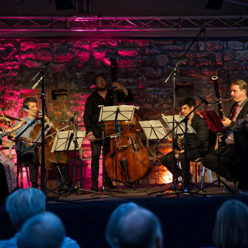 Ein musikalisches Ensemble spielt auf einer Bühne mit leuchtenden Lichtern. Im Hintergrund sind Steinmauern zu sehen, während das Publikum aufmerksam zuhört.