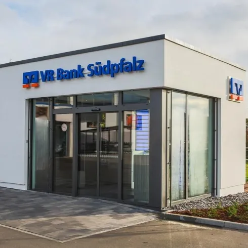 Eine moderne Bankfiliale mit großen Fenstern und einem klaren, einladenden Design. Die Fassade trägt das Logo der VR Bank Südpfalz.