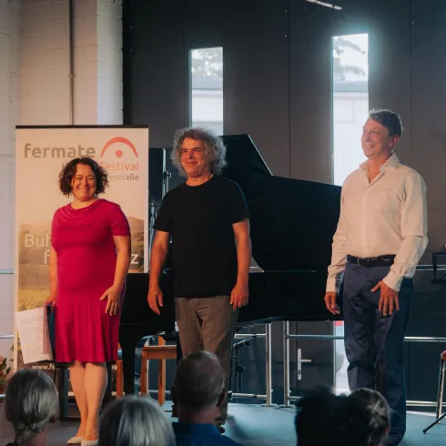 Drei Personen stehen auf einer Bühne und lächeln. Im Hintergrund ist ein Piano und eine entspannte Atmosphäre während einer Veranstaltung zu sehen.