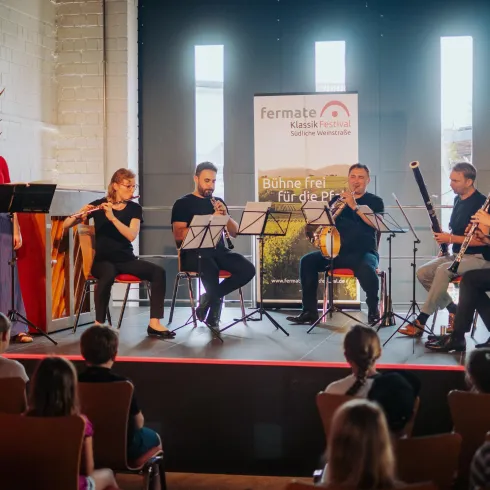 Drei Personen stehen auf einer Bühne und lächeln. Im Hintergrund ist ein Piano und eine entspannte Atmosphäre während einer Veranstaltung zu sehen.