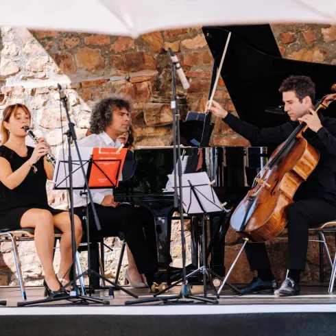 Ein Musiker-Trio spielt auf einer Bühne im Freien. Die Darbietung umfasst Gesang, Klavier und Cello.