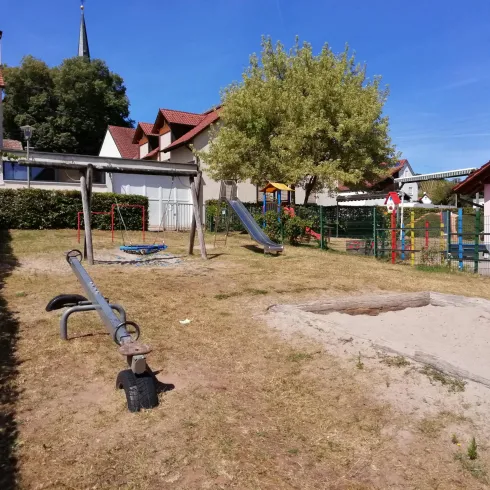 Ein Spielplatz mit Schaukeln und Rutschen in einer ruhigen Nachbarschaft. Im Hintergrund sind einige Häuser und ein großer Baum zu sehen.